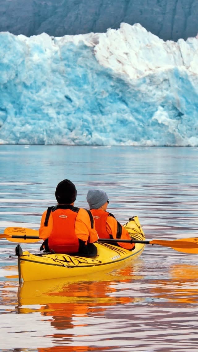 Two kayakers in orange gear exploring Arctic waters near glacier